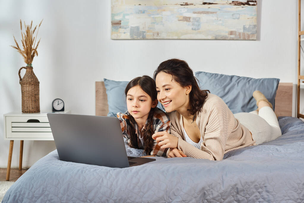 Matka i córka relaksujące się na łóżku, pochłonięte ekranem laptopa. - Zdjęcie, obraz