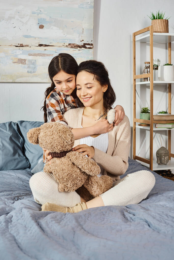 Een moeder en dochter zittend op een bed, knuffelend met een teddybeer in een hartverwarmende omhelzing, genietend van kwaliteitstijd samen. - Foto, afbeelding
