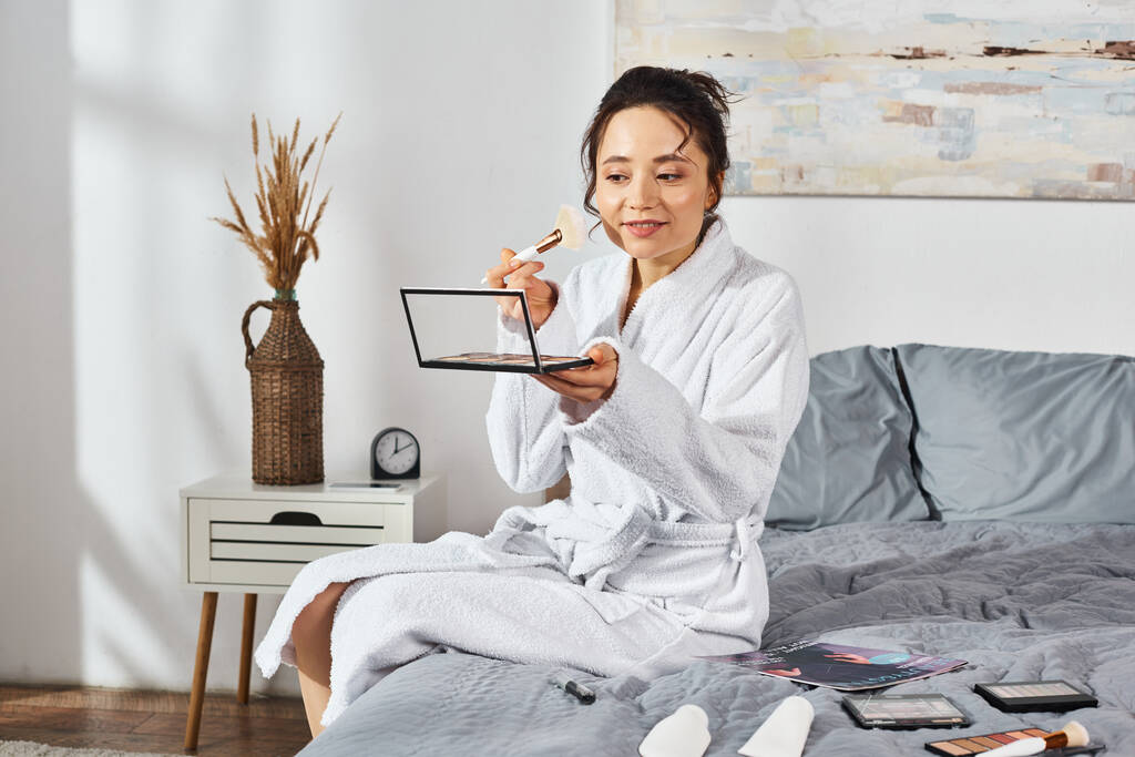 Eine brünette Frau im weißen Bademantel sitzt auf einem Bett mit Lidschatten, umgeben von Kosmetika und schminkt sich morgens.. - Foto, Bild