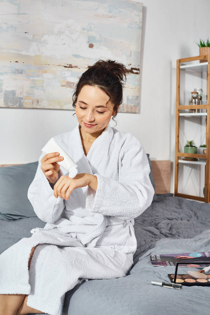 A brunette woman sitting on a bed in a white bathrobe, applying cream in the morning. - Photo, Image