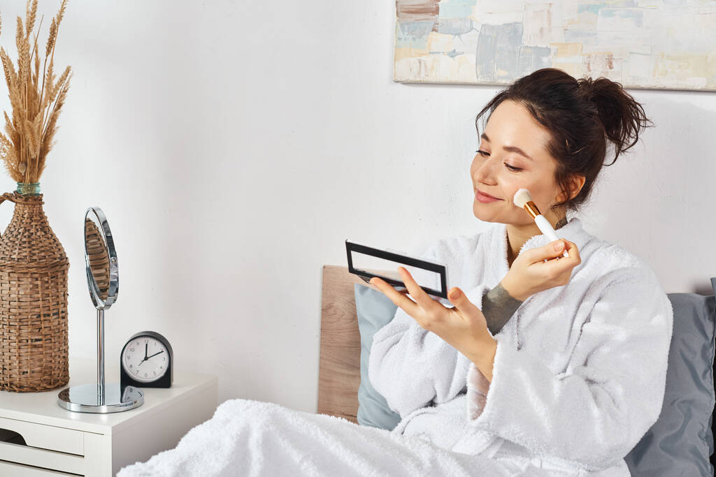 Una donna bruna in accappatoio bianco si siede su un letto, applica il trucco al mattino. - Foto, immagini