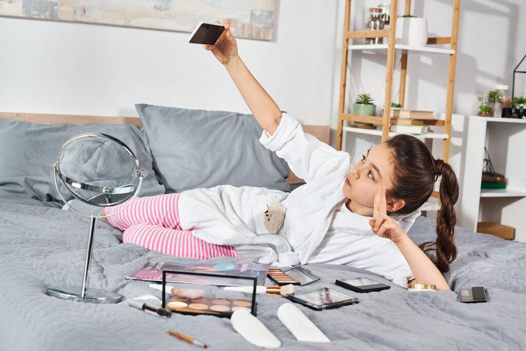 Egy barna tinédzser lány fehér köntösben fekszik egy ágyon, mobiltelefonnal a kezében, elmerülve a gondolataiban.. - Fotó, kép
