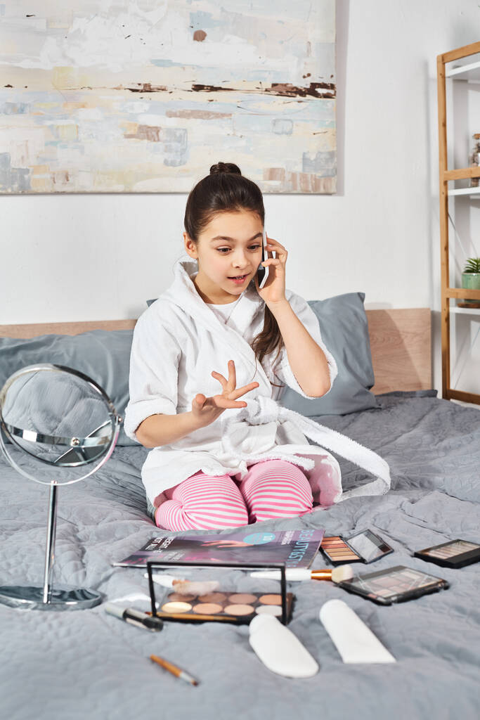 Ein junges Mädchen im weißen Bademantel sitzt auf einem Bett und plaudert mit ihrem Handy. - Foto, Bild