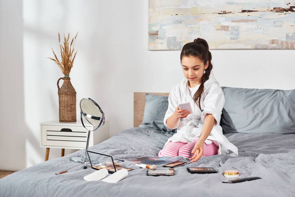 Une jeune fille brune en peignoir blanc s'assoit sur un lit, absorbée dans son téléphone portable. - Photo, image