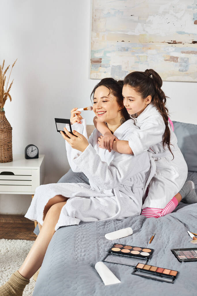 Een brunette moeder en haar dochter in witte badjassen zitten samen op een gezellig bed, het delen van een speciaal moment. - Foto, afbeelding