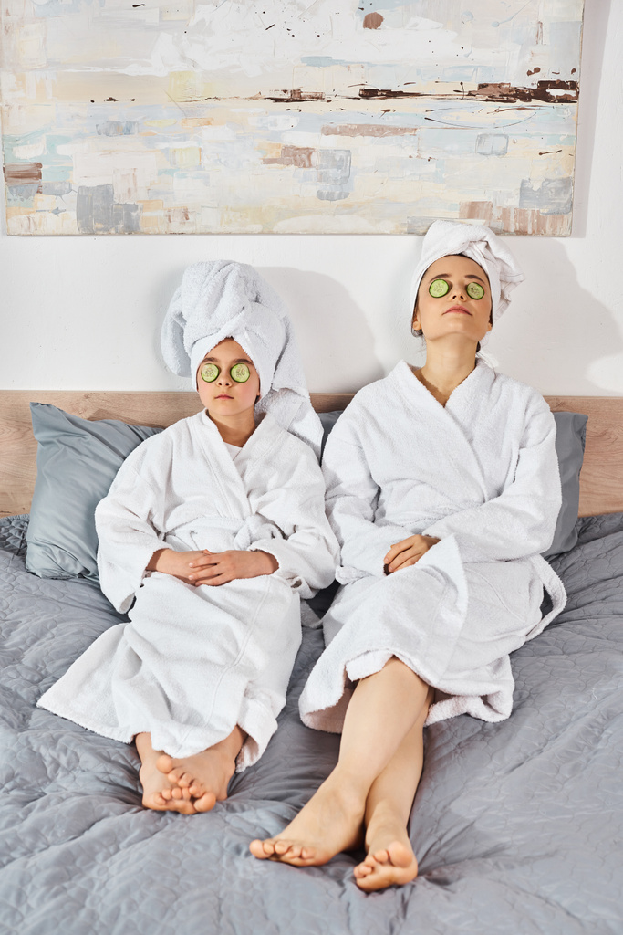 Mutter und Tochter in weißen Bademänteln auf dem Bett sitzend, Handtücher um den Kopf gewickelt, teilen einen schönen Moment. - Foto, Bild