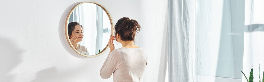 Une femme brune se tient devant un miroir, appliquant de la crème visage dans une salle de bain moderne. - Photo, image