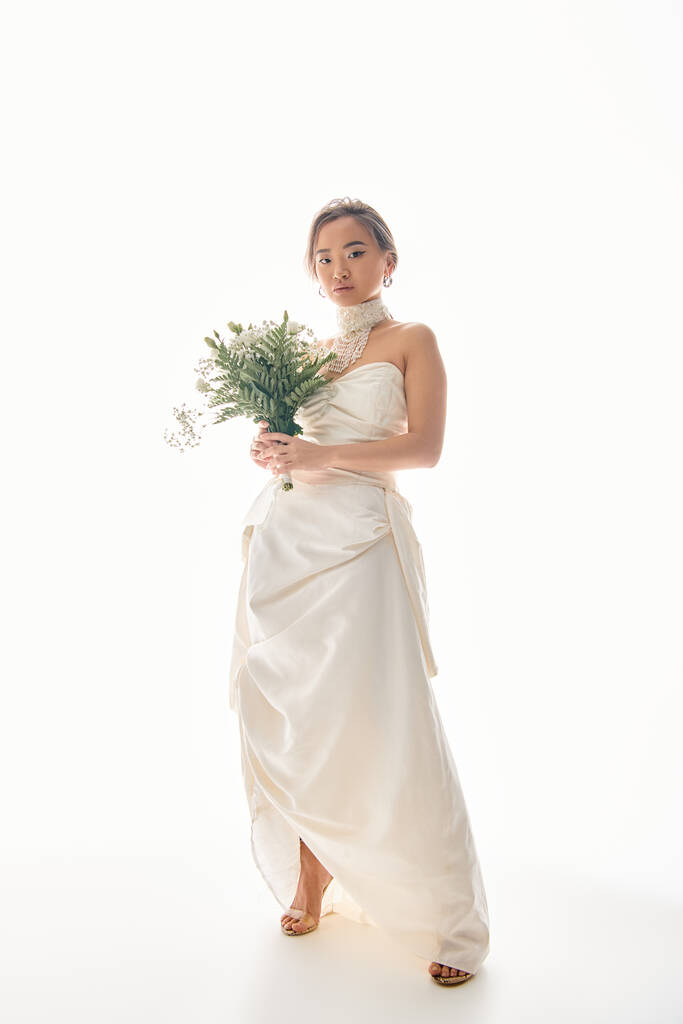 витончена азіатська молода жінка в білій сукні позує на букет квітів на світлому фоні - Фото, зображення