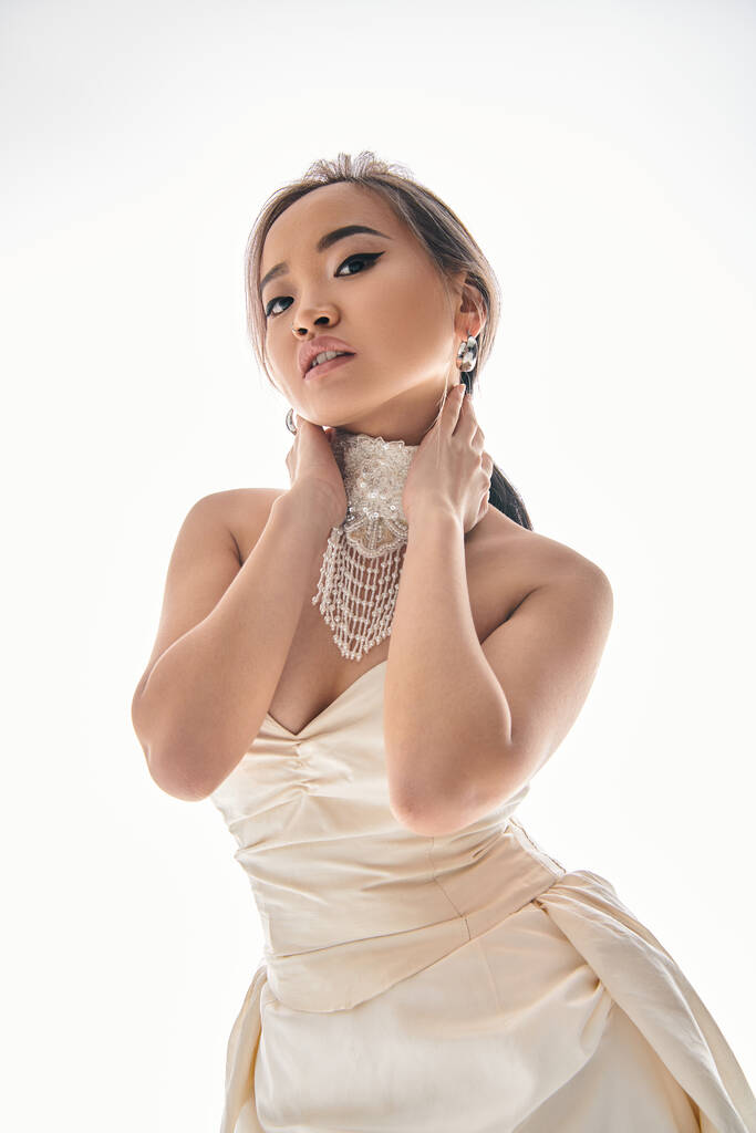 elegante asiático jovem mulher no branco vestido olhar para câmera e abraço pescoço com as mãos no luz fundo - Foto, Imagem