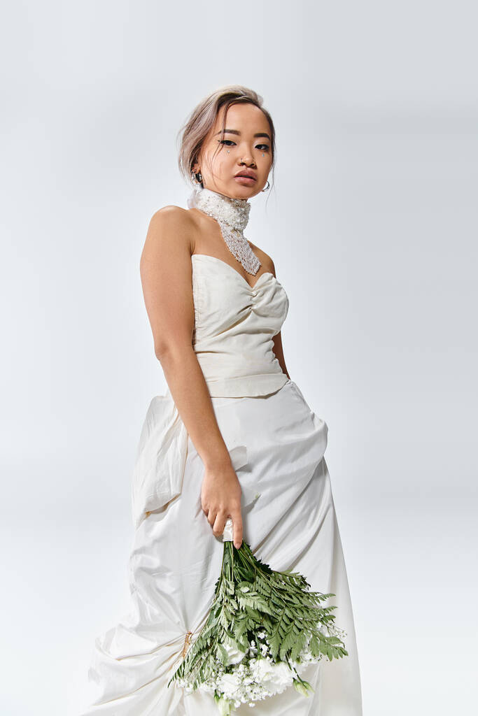 uroczy Azji młoda kobieta w biały elegancki strój pozowanie z kwiaty bukiet na lekkim tle - Zdjęcie, obraz