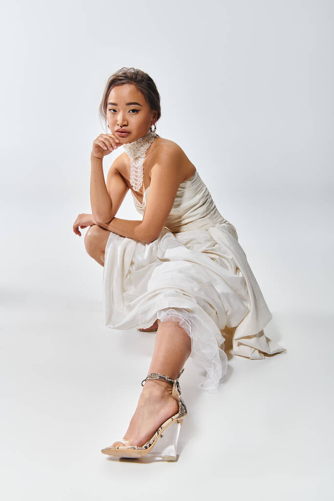 aantrekkelijke aziatische jonge vrouw in wit stijlvolle jurk hurkt naar beneden en gestrekt been uit in de voorkant - Foto, afbeelding