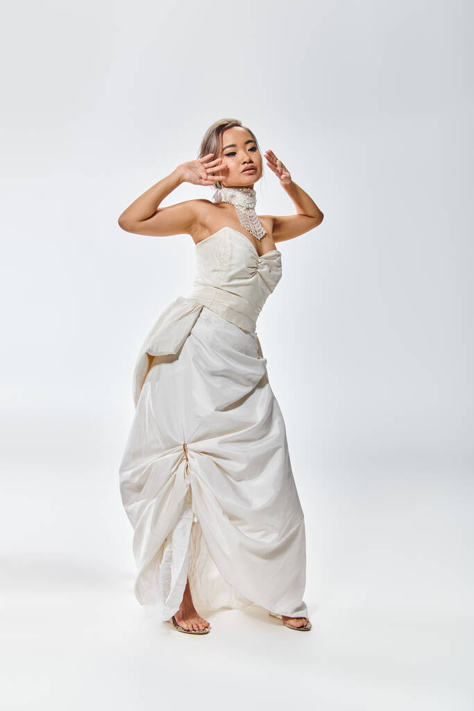 encantador asiático noiva em branco vestido de graça posando com as mãos perto do rosto no fundo de luz - Foto, Imagem