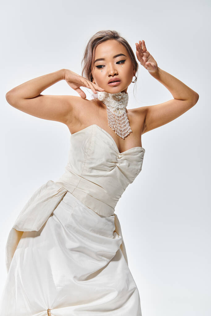 елегантна азіатська жінка в білій витонченій сукні позує руками біля обличчя і дивиться вниз - Фото, зображення