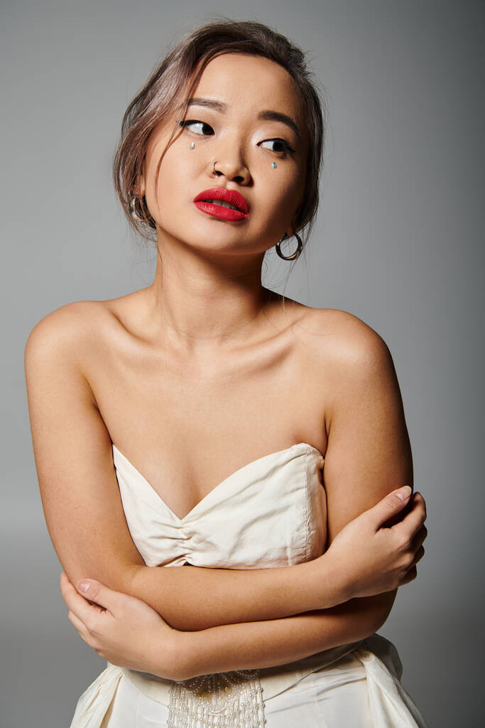 charmante asiatique jeune femme avec rouge à lèvres se câliner et regarder de côté sur fond gris - Photo, image