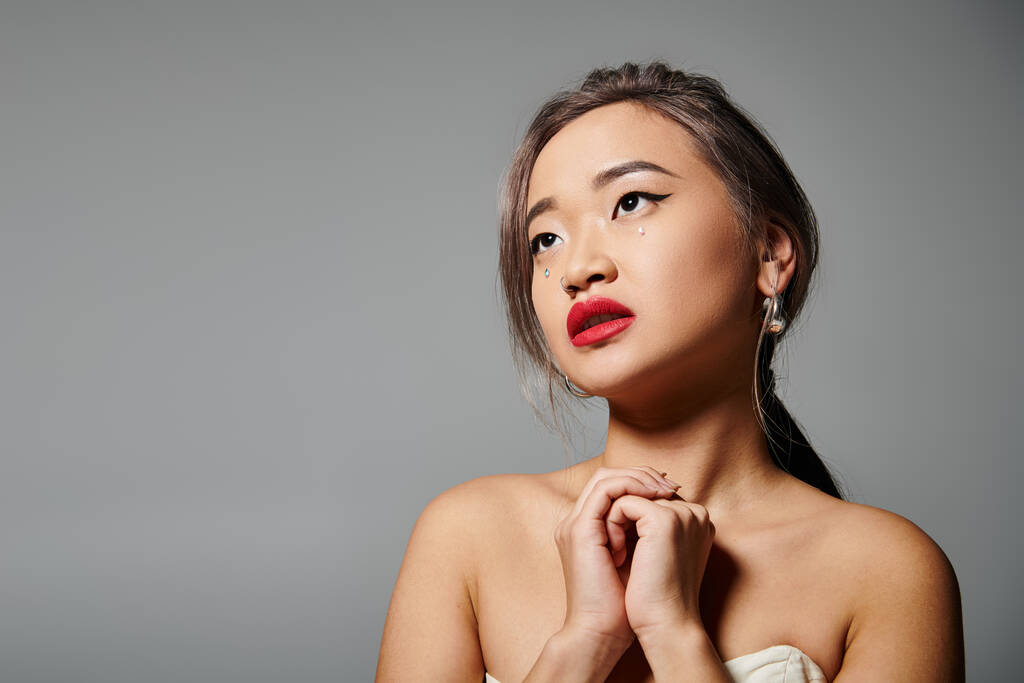 attractive asian woman with red lips folding hands and looking to up against grey background - Photo, Image