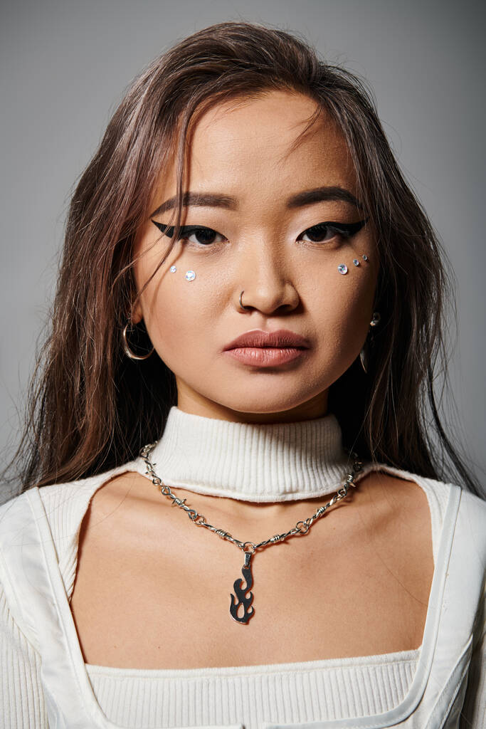 hermosa mujer joven asiática con atrevido maquillaje y collar mirando a la cámara sobre fondo gris - Foto, imagen