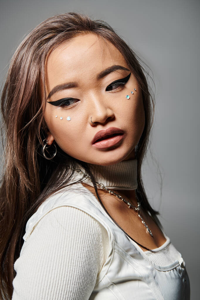 sedutor asiático jovem mulher com pesado maquiagem olhando para trás contra cinza fundo - Foto, Imagem