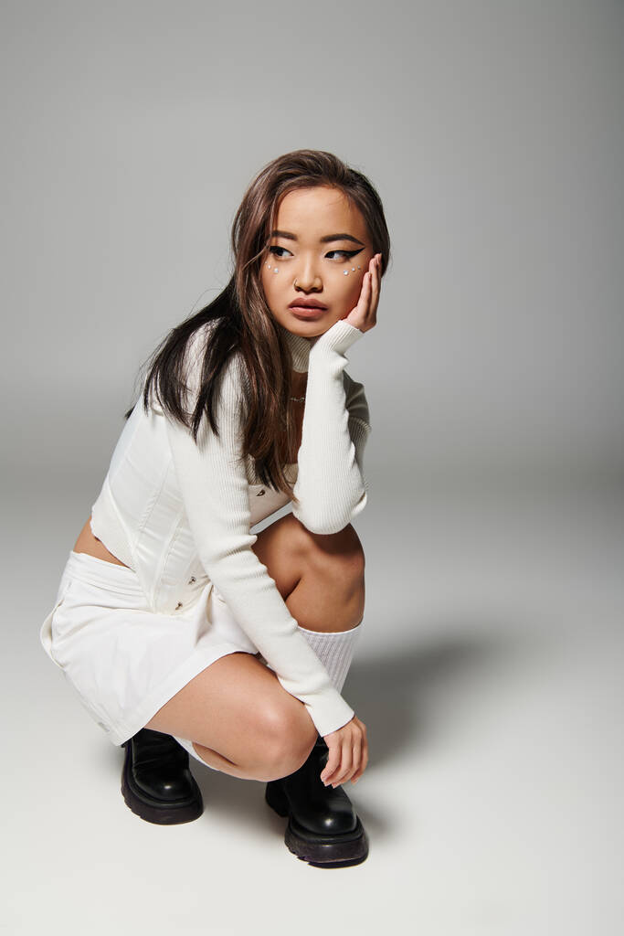 attractive asian woman in her 20s with heavy makeup sideways crouched down on grey background - Photo, Image