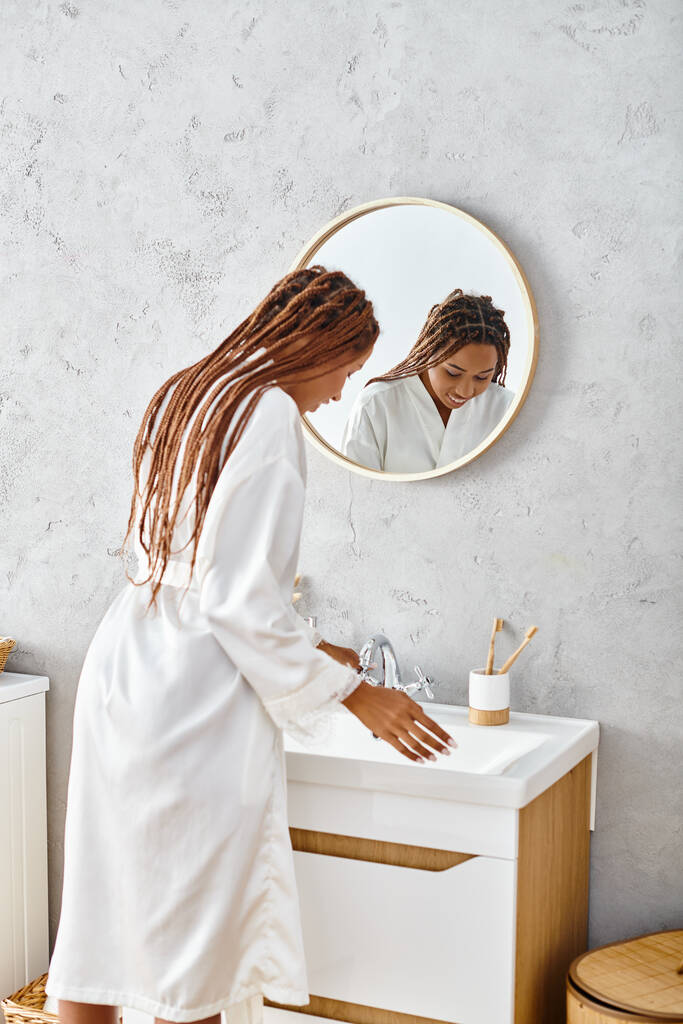 Uma mulher afro-americana com tranças afro fica em seu banheiro moderno, engajando-se em rituais de beleza e higiene.. - Foto, Imagem