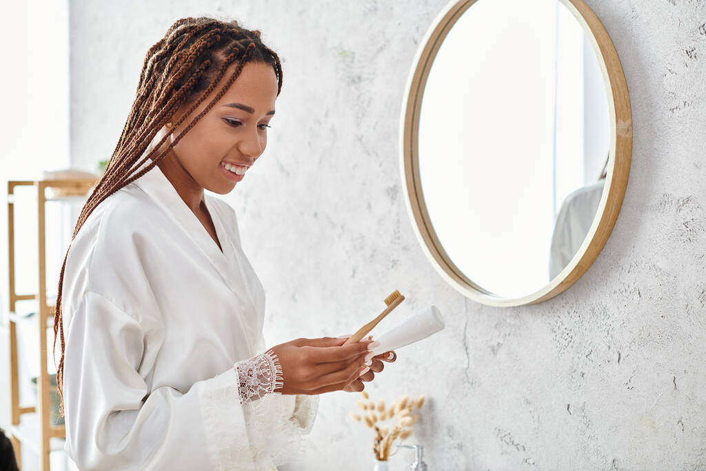 Una donna afro-americana con trecce afro si trova in un bagno moderno, con una spazzola in mano mentre indossa un accappatoio. - Foto, immagini