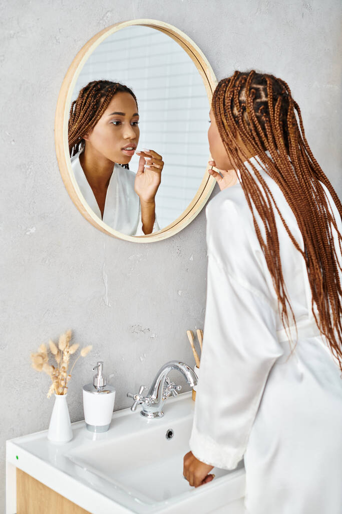 Афроамериканка с афрокосичками чистит зубы в современном зеркале в ванной, надевая халат.. - Фото, изображение