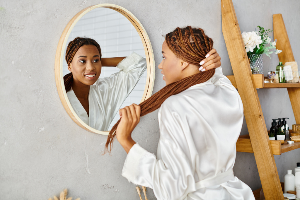 Стильная афроамериканка с афрокосичками расчесывает волосы перед зеркалом в современной ванной. - Фото, изображение