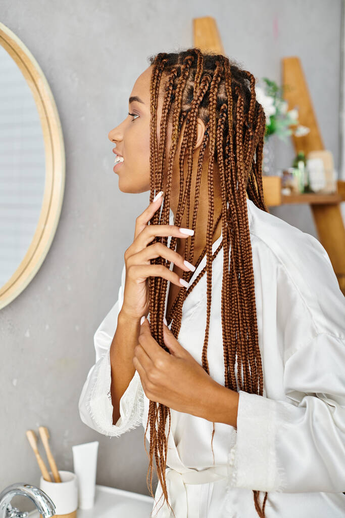 Una mujer afroamericana con trenzas afro cepillándose el pelo en un baño moderno mientras usa una bata de baño. - Foto, imagen