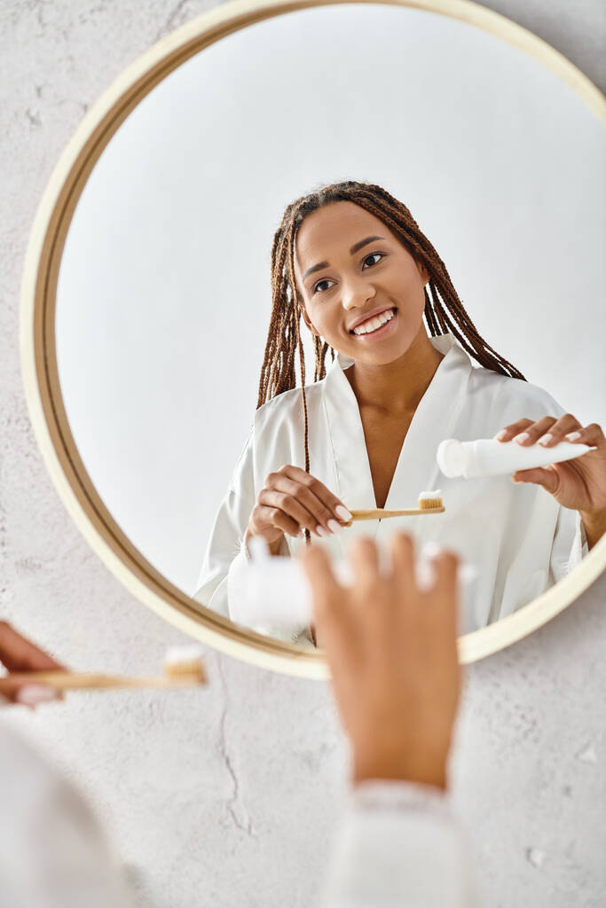 Egy afro-copfos afro-amerikai nő fürdőköpenyben, aki a tükör előtt mossa a fogát egy modern fürdőszobában.. - Fotó, kép