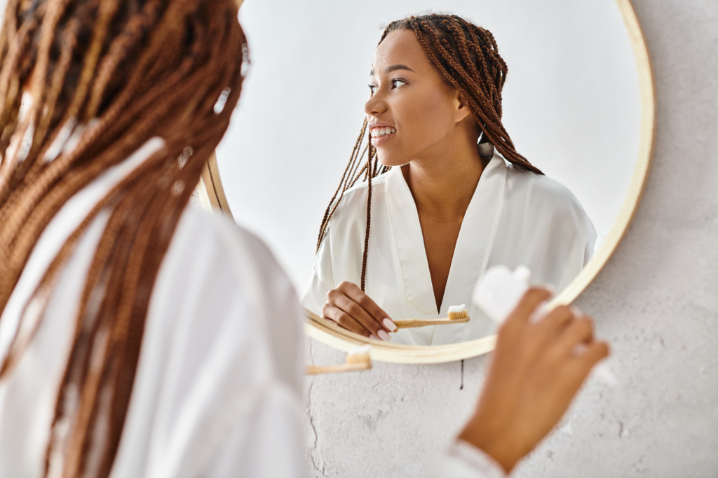 Una mujer afroamericana en albornoz con trenzas afro se cepilla los dientes frente a un espejo en un baño moderno. - Foto, Imagen