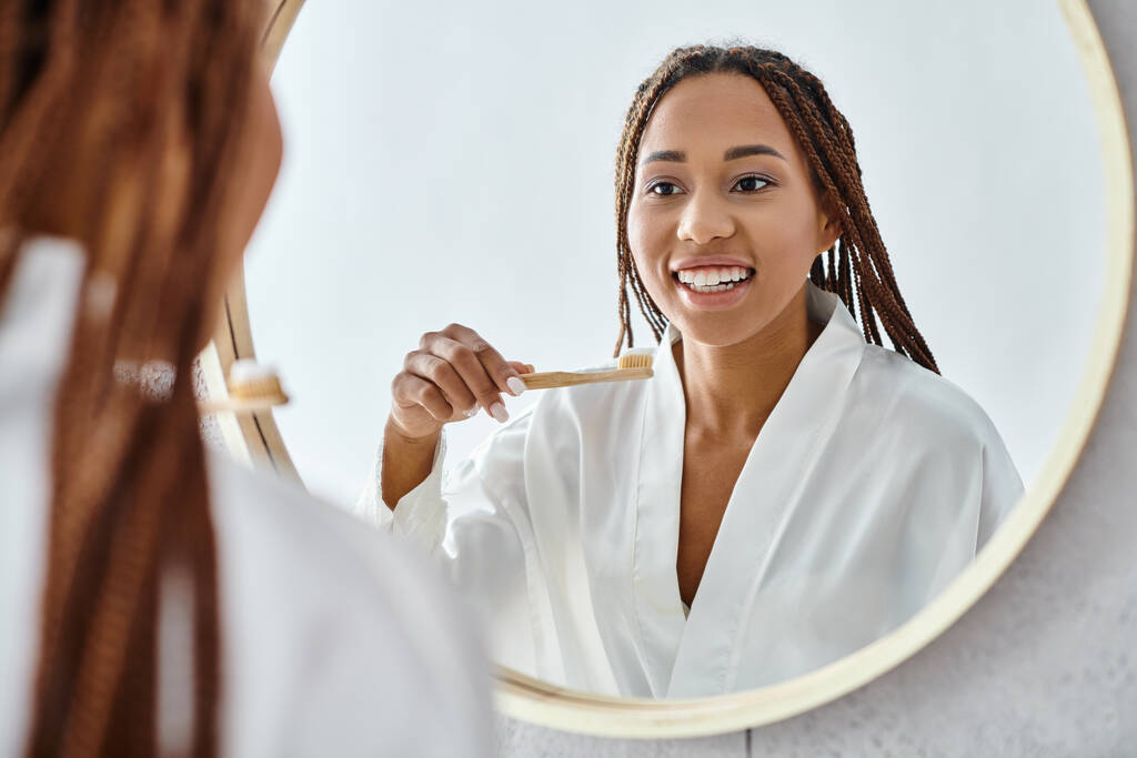 Uma mulher afro-americana com tranças afro, em um manto de banho, escovando os dentes em um espelho de banheiro moderno. - Foto, Imagem