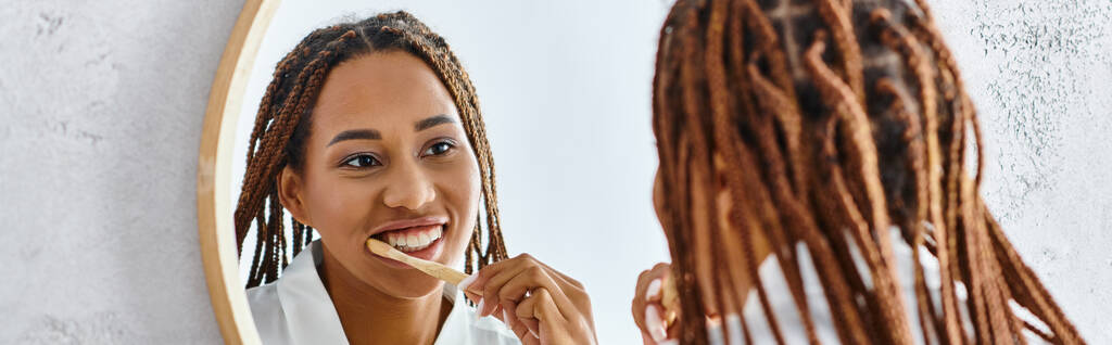 Een Afro-Amerikaanse vrouw met afro vlechten in een badjas poetst haar tanden in een moderne badkamer terwijl ze kijkt naar haar reflectie in de spiegel. - Foto, afbeelding