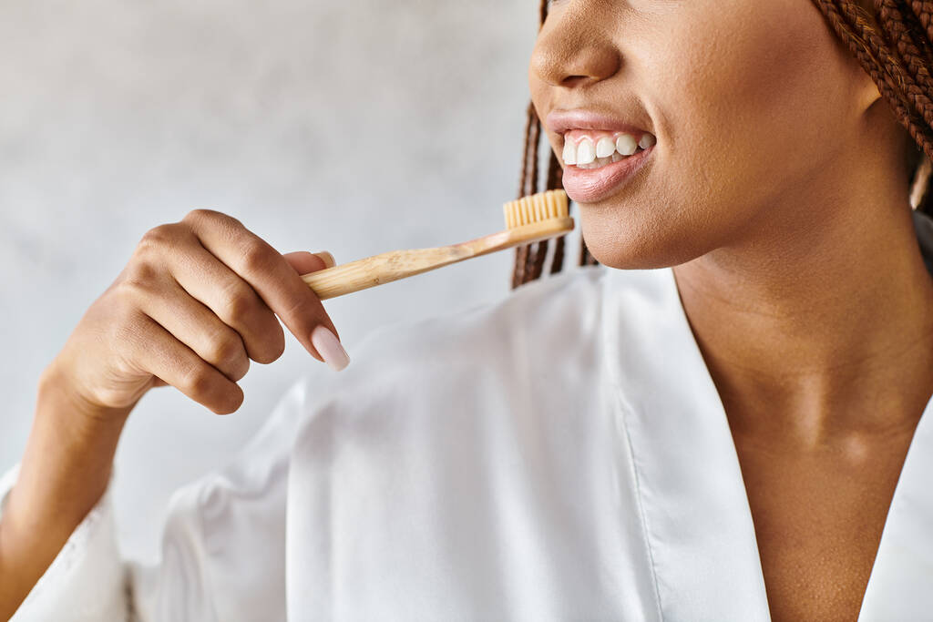 Afroamerykanka z afro warkoczami, w szlafroku, myjąca zęby drewnianą szczoteczką do zębów w nowoczesnej łazience. - Zdjęcie, obraz