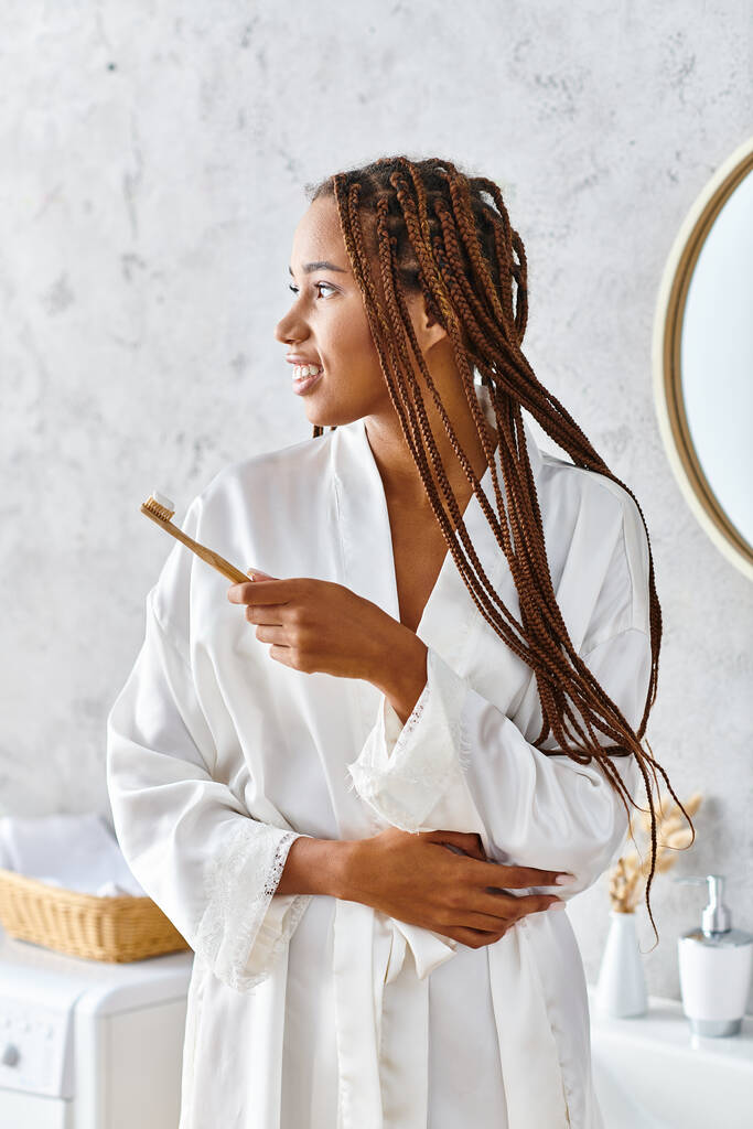 Μια Αφρο-Αμερικανίδα με ράστα, ντυμένη με μπουρνούζι, βουρτσίζοντας τα δόντια της στο μοντέρνο μπάνιο της.. - Φωτογραφία, εικόνα