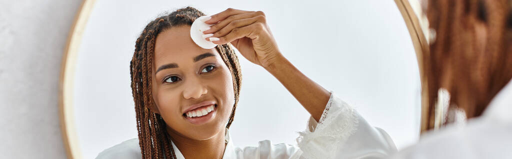 Африканська американка з афро косами, використовуючи бавовняну подушку з тонером перед дзеркалом у сучасній ванній кімнаті, орієнтуючись на красу та гігієну. - Фото, зображення
