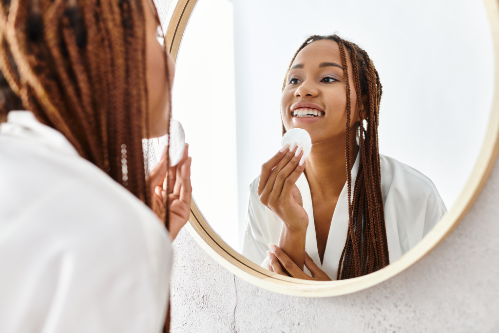 Африканська американська жінка з афро косами в халаті, використовуючи бавовняну подушку перед дзеркалом у сучасній ванній кімнаті. - Фото, зображення