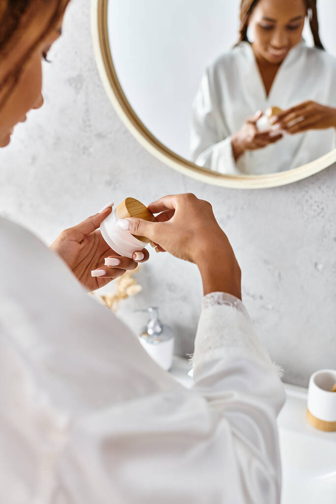 Афроамериканка с афрокосичками в халате держит банку красоты со сливками перед зеркалом в современной ванной комнате. - Фото, изображение
