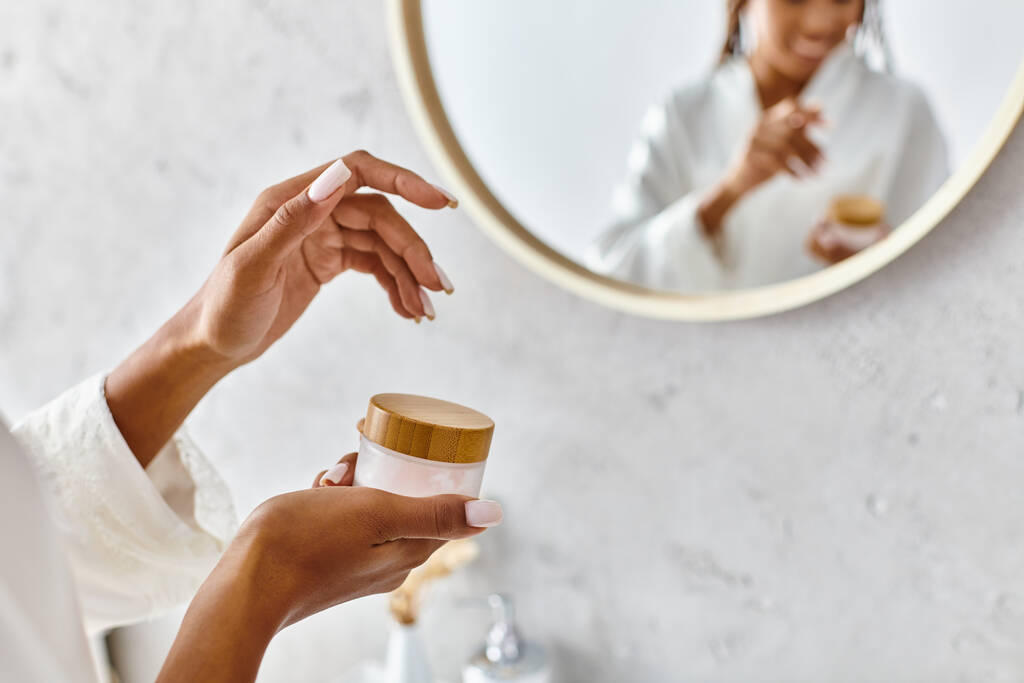 Egy afro-amerikai nő fürdőköpenyben, kezében egy üveg tejszínnel a tükör előtt a modern fürdőszobájában.. - Fotó, kép