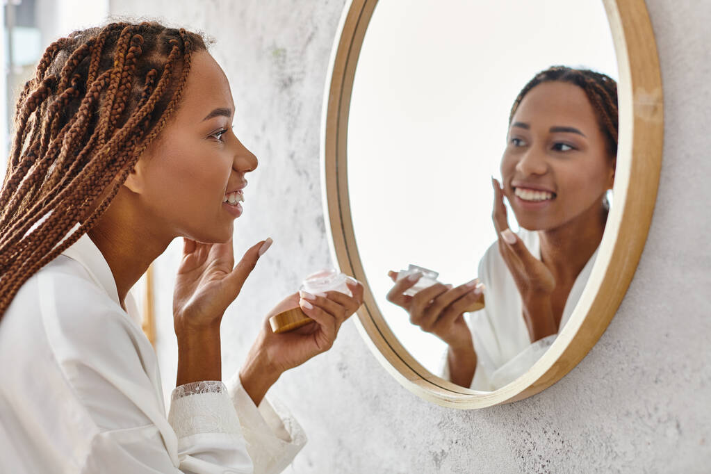 Een Afro-Amerikaanse vrouw met afro vlechten aanbrengen crème in een moderne badkamer terwijl het dragen van een badjas. - Foto, afbeelding