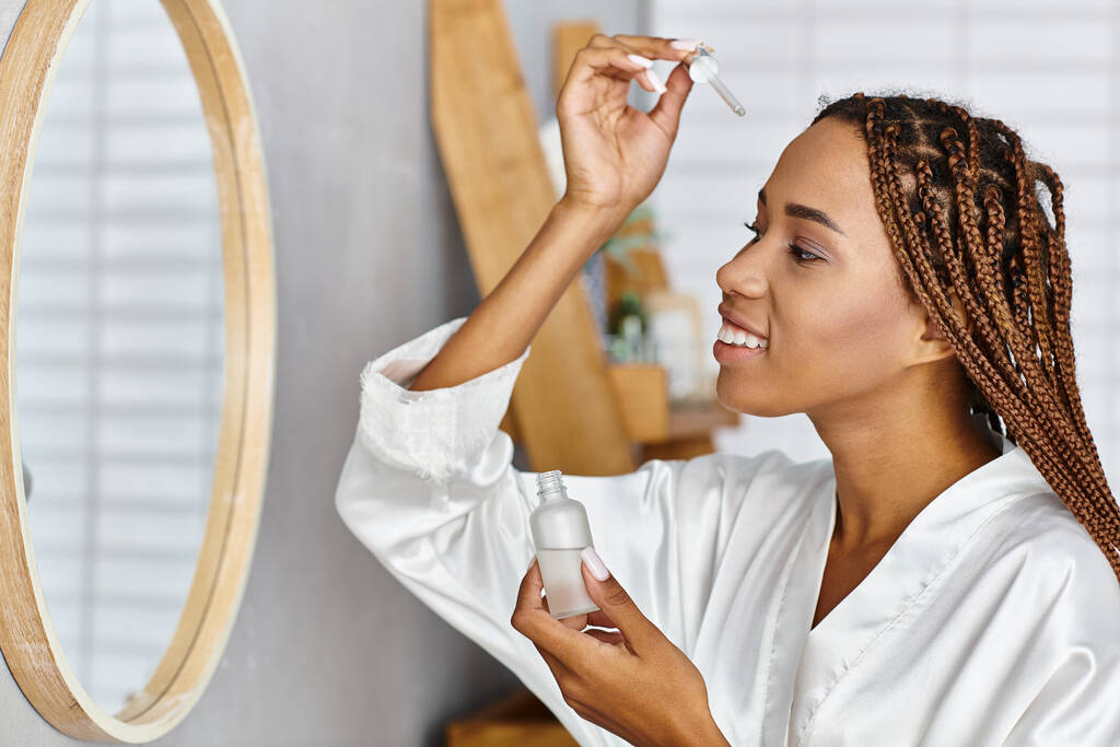 Afroamerykanka z afro warkoczami trzymająca butelkę z serum przed nowoczesnym lustrem w łazience. Piękno i higiena rutyna. - Zdjęcie, obraz