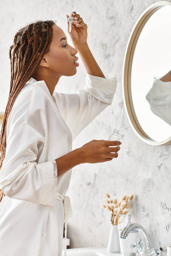 Une Afro-Américaine avec des tresses afro tenant une bouteille avec du sérum dans une salle de bain moderne tout en portant un peignoir de bain. - Photo, image