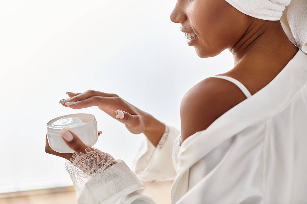 Афроамериканка в белом платье мирно держит банку крема в современной ванной комнате, олицетворяя красоту и элегантность. - Фото, изображение