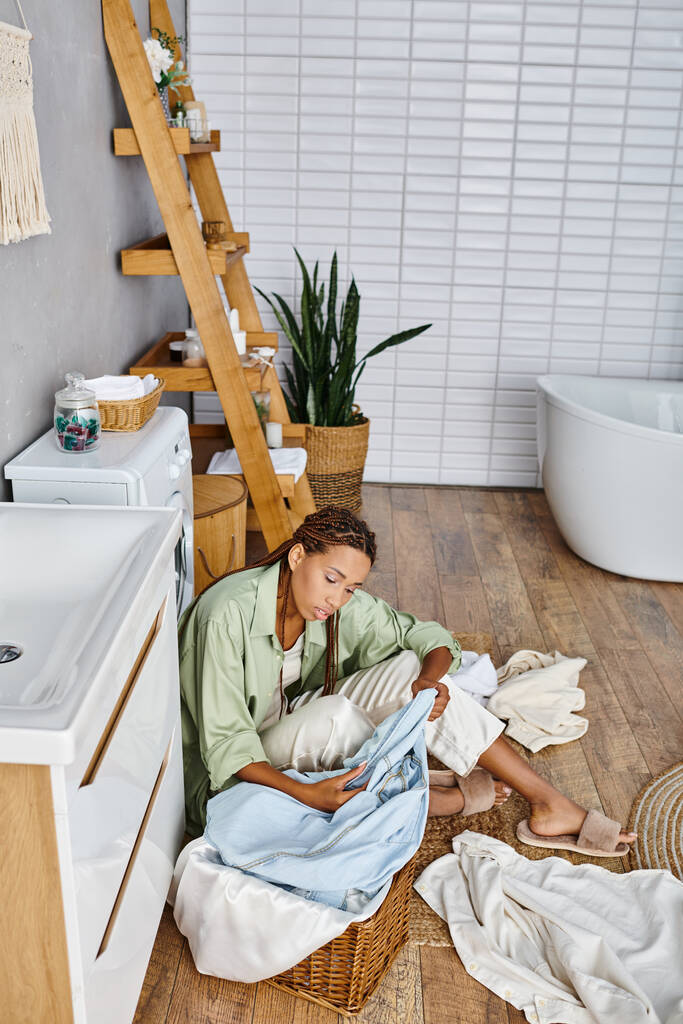 Eine afroamerikanische Frau mit Afro-Zöpfen sitzt auf dem Badezimmerboden, umgeben von Wäsche, während sie Hausarbeit erledigt. - Foto, Bild