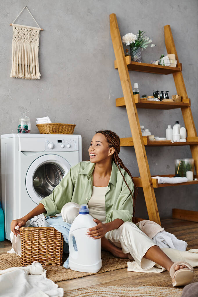 Een Afro-Amerikaanse vrouw met afro vlechten rustig zit op de vloer naast een wasmachine, het doen van de was in een badkamer. - Foto, afbeelding