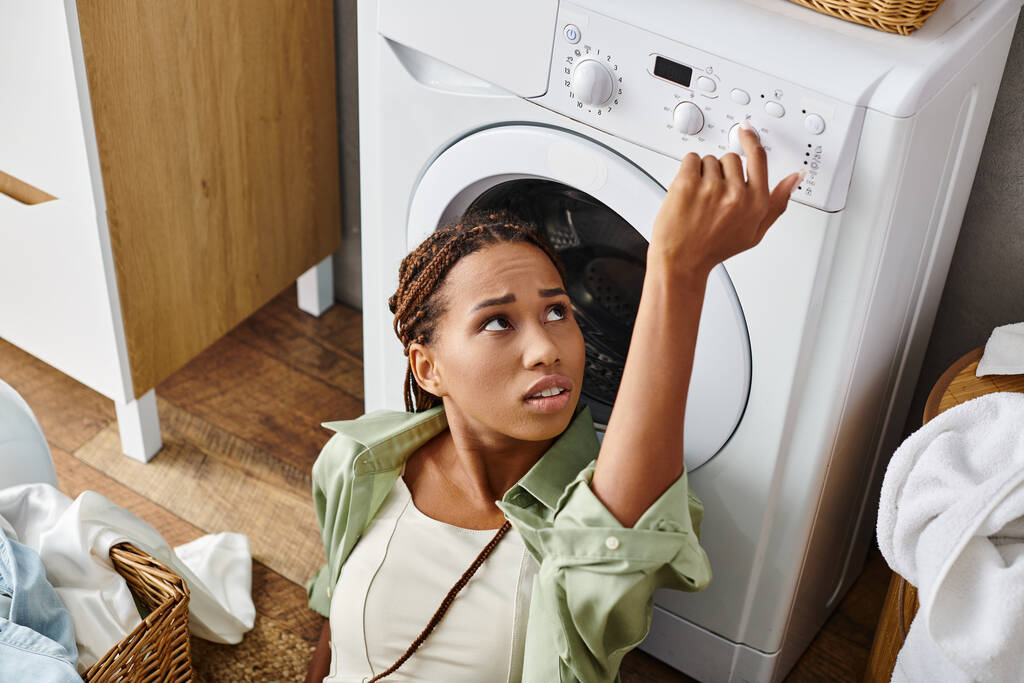 Una mujer afroamericana con trenzas afro mira hacia arriba a una secadora mientras hace la colada en un baño. - Foto, imagen