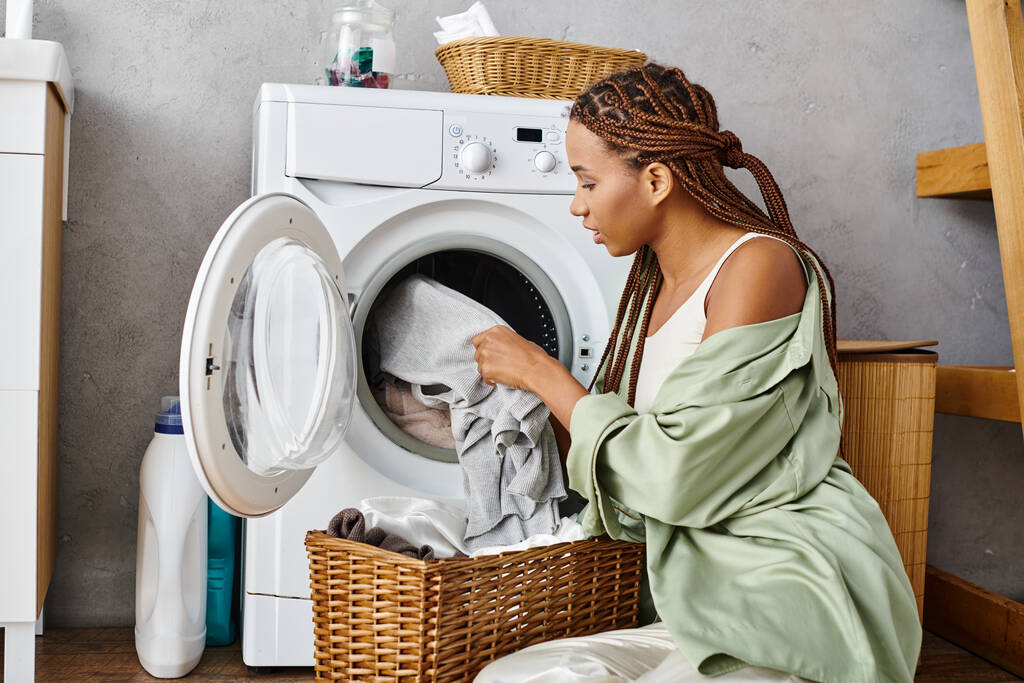 Afro örgülü Afrikalı bir kadın çamaşır makinesinin yanında oturmuş banyoda çamaşır yıkıyor.. - Fotoğraf, Görsel