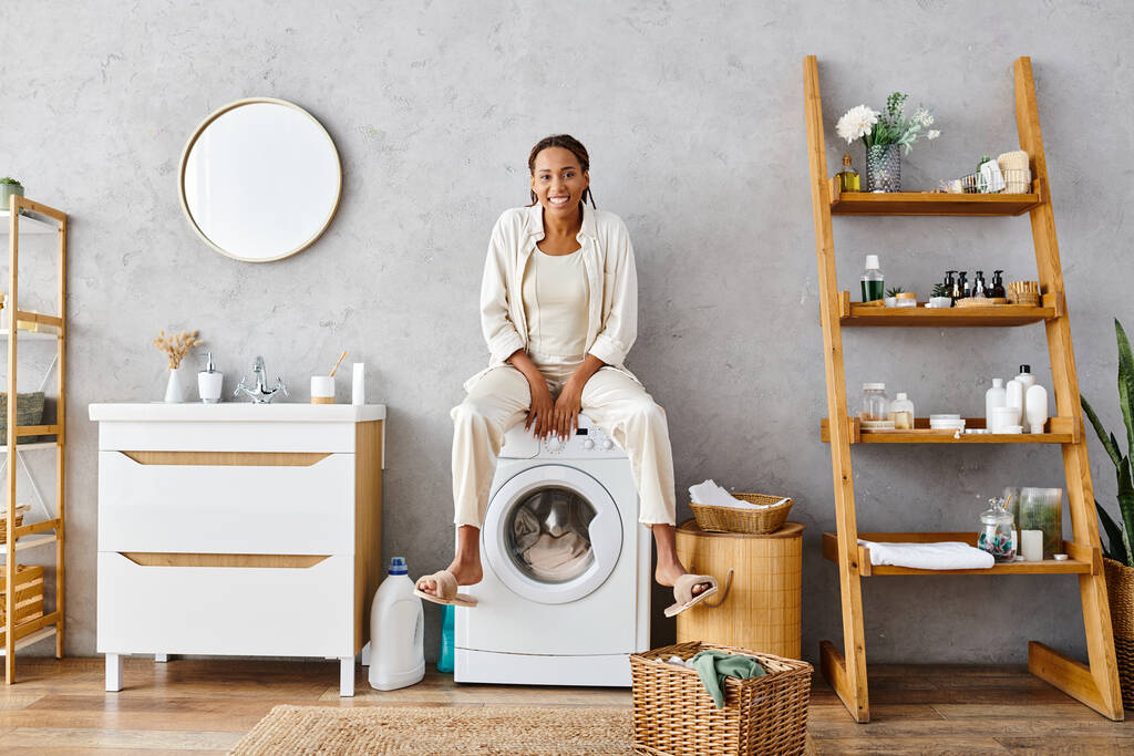 Афроамериканка с афрокосичками сидит на стиральной машине и стирает бельё в ванной комнате.. - Фото, изображение