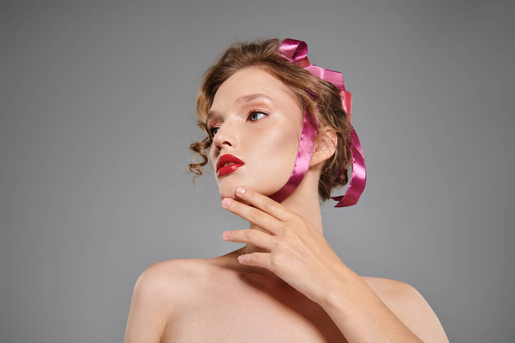 Νεαρή γυναίκα αποπνέει κλασική ομορφιά καθώς ποζάρει με ένα ροζ φιόγκο στο κεφάλι της σε ένα στούντιο σε γκρι φόντο. - Φωτογραφία, εικόνα