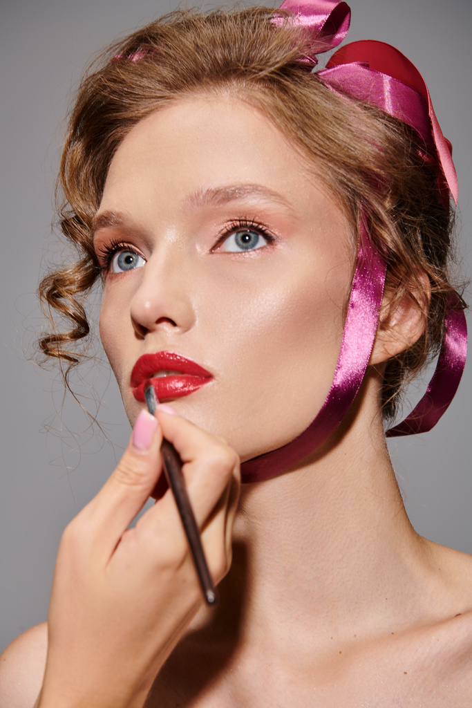 Una belleza clásica, una joven posa elegantemente con un lazo rosa posado sobre su cabeza en un estudio sobre un fondo gris. - Foto, Imagen