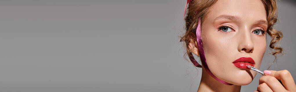 Une jeune femme en studio applique gracieusement du rouge à lèvres sur ses lèvres avec un pinceau, exsudant une beauté classique. - Photo, image