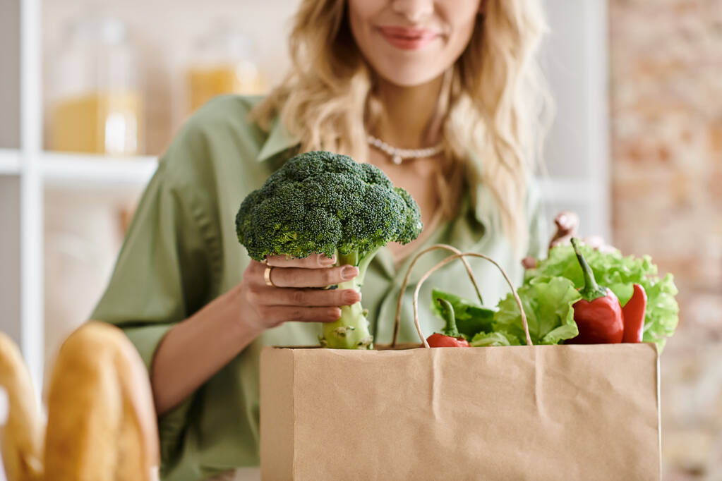 Una mujer sosteniendo una bolsa de papel llena de varias verduras frescas en una cocina. - Foto, Imagen